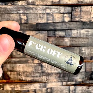 May include: A small, brown glass roller bottle with a white label that reads "F*CK OFF I AM SHIELDED FROM NEGATIVITY I AM A POWERFUL SOURCE OF LIGHT". The label also features a small graphic of a person sitting in a meditative pose.
