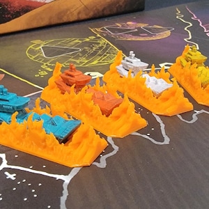 May include: A set of six miniature spaceships on fire, each with a different color hull: blue, orange, white, yellow, red, and teal. The spaceships are on a dark brown surface with white markings.