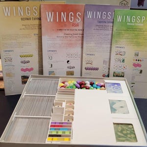 May include: An open board game box with various components, including cards, colorful game pieces, and expansion packs. The expansion packs are labeled "Wings" with different regional themes: Oceania, Asia, European, and Americas.