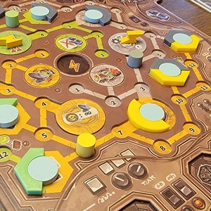 May include: A brown board game with yellow, green, and blue game pieces. The board has a grid pattern with numbers 1 through 16. There are also several small, round, gray pieces on the board.