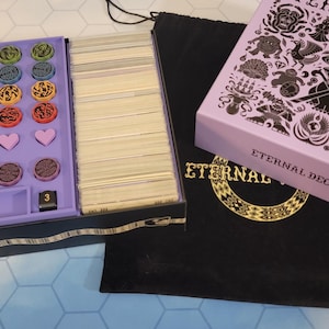 May include: A board game set with a purple box containing game pieces, a stack of cards, and a pink box with the words "Eternal Decks". A black bag with gold lettering and a book with the words "Eternal Decks" are also present.