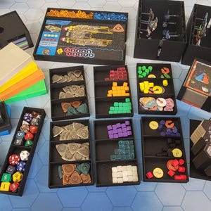 May include: A collection of board game components, including dice, tokens, cards, and miniatures, organized in black trays and boxes. The game box features a spaceship graphic. Various colored game pieces are visible, suggesting a strategy or adventure game.