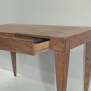 Op de afbeelding: Een houten bureau met een gedeeltelijk open lade. Het bureau is gemaakt van lichtbruin hout en heeft een rechthoekige bovenkant. De poten zijn taps toelopend en de lade is zichtbaar.