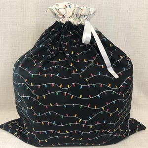 May include: A large black drawstring bag with a repeating pattern of colorful Christmas lights. The top of the bag is white with a similar light pattern and a white ribbon. The bag is made of fabric.