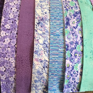 May include: A collection of 12 fabric strips in various shades of purple, blue, and green. The strips feature different patterns, including floral, geometric, and abstract designs.