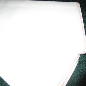 Könnte beinhalten: Ein weißes Baumwoll-Geschirrtuch mit einem roten, gesteppten Rand. Das Tuch ist in der Hälfte gefaltet und liegt auf einer grünen Oberfläche.