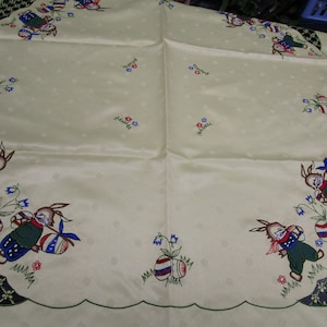 Könnte beinhalten: Eine weiße Tischdecke mit einem gewellten Rand und einem gestickten Muster von Hasen, die Ostereier halten. Die Hasen tragen blaue und grüne Kleidung und die Eier sind mit Rot, Blau und Weiß verziert.