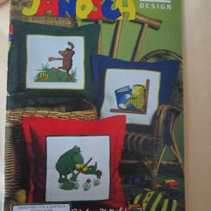 Könnte beinhalten: Ein Rico Design Janosch Band 22 Bastelheft mit drei quadratischen Kissen mit Kreuzstich-Illustrationen eines braunen Bären, eines gelb gestreiften Tigers und eines grünen Frosches. Das Cover zeigt den Text Janosch, Rico Design und Stich-Kollektion.