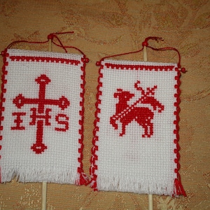 Könnte beinhalten: Zwei kleine, weiß bestickte Banner mit roten Akzenten. Ein Banner zeigt ein rotes Kreuz mit den Buchstaben IHS, das andere ein rotes Lamm, das einen Stab hält. Beide sind an Holzstäben befestigt.