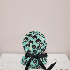 May include: A teal blue round gift box with a black ribbon. The box is decorated with a pattern of black jack-o'-lanterns and ghosts.