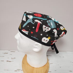 May include: Black scrub hat with a fun dental themed print featuring cartoon teeth, toothbrushes, floss, and other dental tools. The hat has a black tie.