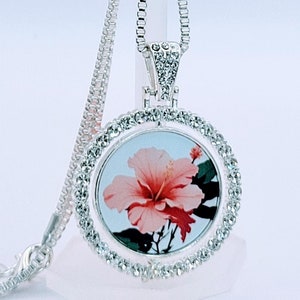 May include: A silver necklace with a round pendant featuring a pink hibiscus flower surrounded by clear rhinestones.