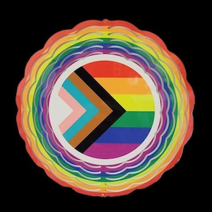 May include: A circular, multi-colored, layered design with a rainbow flag in the center. The flag has a black and brown chevron on a white background, with the traditional rainbow colors below.