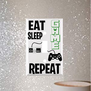 May include: White light switch cover with black and green text and graphics. The text reads "Eat Sleep Game Repeat" with a video game controller and a retro video game console graphic.