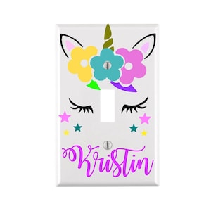May include: White light switch cover with a unicorn design. The unicorn has a yellow, blue, and pink flower crown, closed eyes, and a gold horn. The name "Kristin" is written in purple below the unicorn.