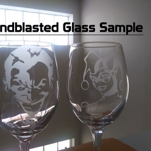 May include: Two clear glass wine glasses with sandblasted designs. The glass on the left features a white silhouette of the Joker, while the glass on the right features a white silhouette of Harley Quinn.