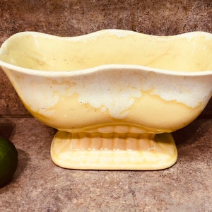 May include: A yellow ceramic planter with a scalloped rim and a pedestal base. The planter has a mottled glaze with white and yellow tones.