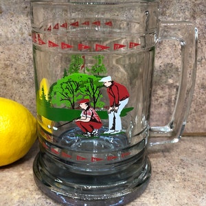 May include: A clear glass golf-themed beer mug with a red, white, and green illustration of two golfers on a green. The mug has a handle and a base with red flags around the rim.