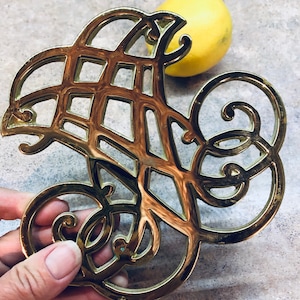 May include: Gold-toned metal hot pad with an intricate, ornate design. The pad is shaped like a stylized letter S.