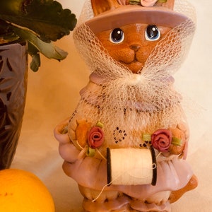 May include: A ceramic cat figurine wearing a hat, veil, and dress, holding a spool of thread. The cat is brown with blue eyes, and the outfit is a light purple with pink roses. The figurine has a vintage, handcrafted appearance.