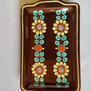 May include: A brown ceramic rectangular dish with a decorative floral design. The dish has a yellow, orange, and green floral pattern on a brown background.
