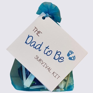 May include: A blue mesh gift bag tied with a blue ribbon, containing small items. A white tag is attached, with the words "The Dad to Be Survival Kit" in blue script and two blue baby footprints.