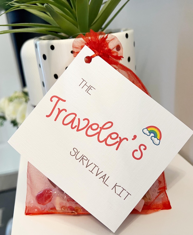 May include: A white tag with the text "The Traveler's Survival Kit" in red script, featuring a rainbow graphic. The tag is attached to a red mesh bag containing small items. The bag is tied with a red ribbon and sits in front of a black and white polka dot pot with a green plant.