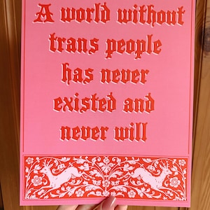 Puede incluir: Un póster rosa con un borde rojo y texto rojo que dice "Un mundo sin personas trans nunca ha existido y nunca existirá". El póster tiene un diseño floral rojo y blanco en la parte inferior con dos ciervos blancos.