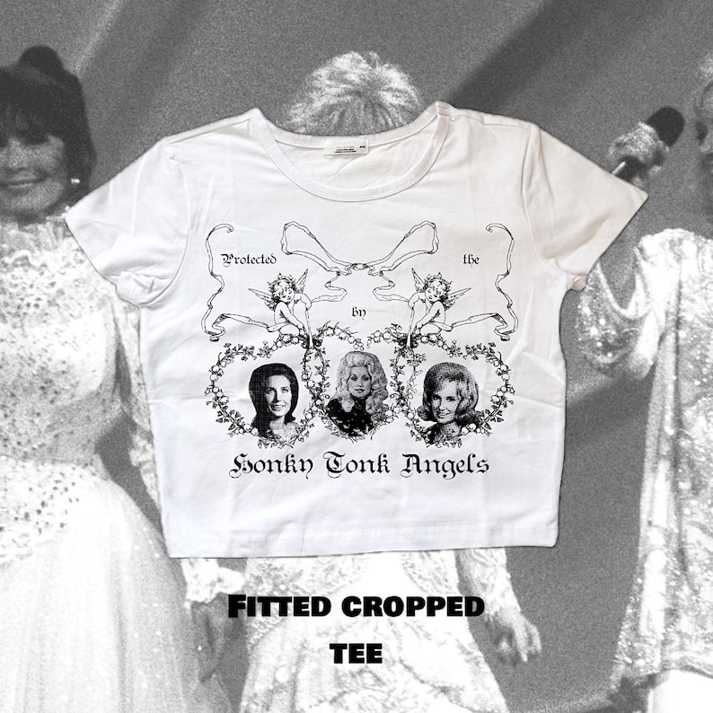Pu&ograve; includere: Maglietta bianca a maniche corte e crop top con una grafica nera di tre donne con ali d'angelo e il testo "Protected by the Honky Tonk Angels". La maglietta ha anche il testo "Fitted Cropped Tee" stampato in basso.