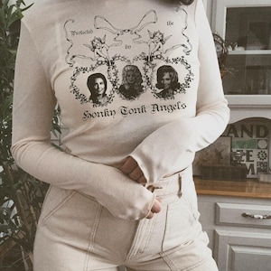 Pu&ograve; includere: Una maglietta bianca a maniche lunghe con una stampa grafica di tre donne in cornici ovali con il testo "Protected by the Honky Tonk Angels".