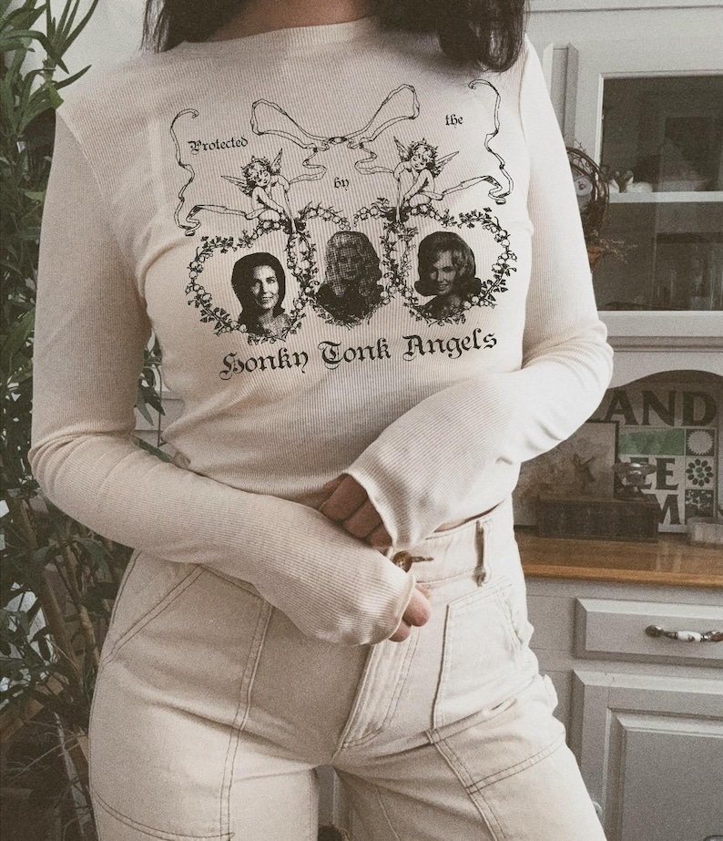 Pu&ograve; includere: Una maglietta bianca a maniche lunghe con una stampa grafica di tre donne in cornici ovali con il testo "Protected by the Honky Tonk Angels".