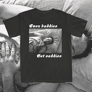 Puede incluir: Camiseta negra con texto blanco que dice "Even baddies get saddies". La camiseta presenta una imagen en blanco y negro de una persona acostada en la cama.