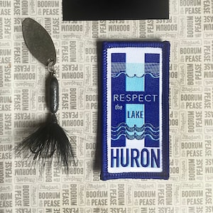 Puede incluir: Un parche azul y blanco con el texto "Respect the Lake Huron" sobre un fondo blanco con un borde azul. El parche está sobre un fondo blanco con un patrón repetido del texto "Boorum & Pease".