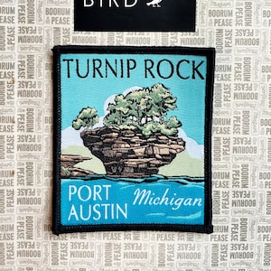 Puede incluir: Un parche bordado azul y negro con el texto "Turnip Rock, Port Austin, Michigan". El parche presenta una ilustración de dibujos animados de una formación rocosa con árboles en la cima, rodeada de agua.