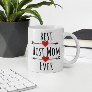 Pode incluir: Caneca de cerâmica branca com o texto "BEST HOST MOM EVER" em preto, com detalhes de corações vermelhos e desenhos de setas. A caneca está sobre uma superfície branca, com um teclado, uma planta e livros ao fundo.