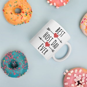 Pode incluir: Caneca de cerâmica branca com texto preto que diz "Best Host Dad Ever" com dois corações vermelhos e flechas.