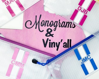 May include: Five clear plastic zippered pouches with different colored stripes and white letters. The pouches are arranged on a pink surface with the text "Monograms & Viny'all" written in black. The pouches are labeled with the initials "MAN", "MRS", "DJB", "CMM", and "JMC".