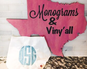 May include: A white bandana with a light blue monogram in the center. The bandana is in front of a pink wooden cutout of the state of Texas with the text "Monograms & Viny'all" in black.