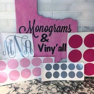 May include: A pink wooden sign with the text "Monograms & Viny'all" in black lettering. There are several sheets of vinyl stickers with various colors and designs, including a monogram, pink circles, and gray circles.