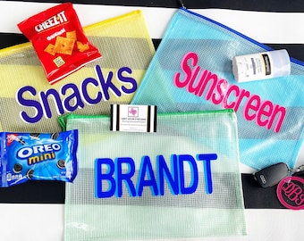 May include: Three clear mesh zippered pouches with different colored stripes. The pouches are labeled "Snacks", "Sunscreen", and "BRANDT". The pouches are filled with various items, including a box of Cheez-It crackers, a box of Oreo mini cookies, and a bottle of sunscreen.