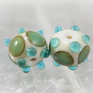 May include: Two handmade glass beads. Each bead is round with a cream-colored base, adorned with teal and green accents. The beads have a series of teal-colored protrusions and green oval shapes. The beads are for jewelry making.