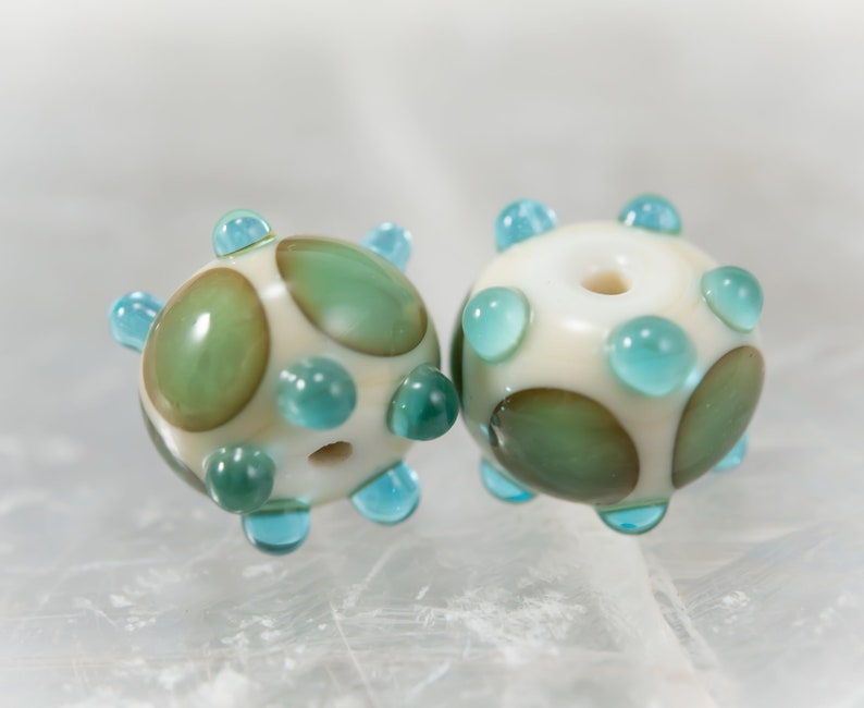 May include: Two handmade glass beads. Each bead is round with a cream-colored base, adorned with teal and green accents. The beads have a series of teal-colored protrusions and green oval shapes. The beads are for jewelry making.