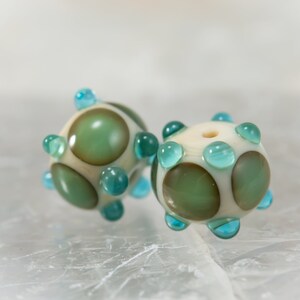 May include: Two handmade glass beads. Each bead is spherical, with a cream-colored base and olive green and teal accents. The beads have a series of teal-colored, rounded protrusions. The beads are likely for jewelry making.