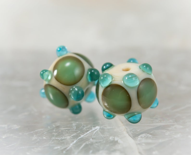 May include: Two handmade glass beads. Each bead is spherical, with a cream-colored base and olive green and teal accents. The beads have a series of teal-colored, rounded protrusions. The beads are likely for jewelry making.