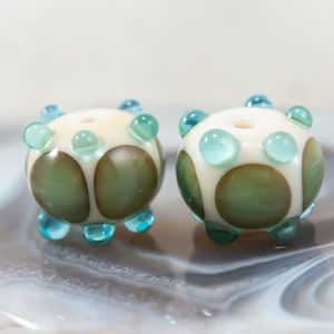 May include: Two handmade glass beads. Each bead is round with a cream-colored base, adorned with green and brown circular accents. They feature turquoise-colored, rounded protrusions. The beads are displayed on a marbled surface.