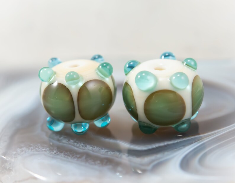 May include: Two handmade glass beads. Each bead is round with a cream-colored base, adorned with green and brown circular accents. They feature turquoise-colored, rounded protrusions. The beads are displayed on a marbled surface.
