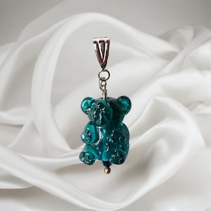 May include: A teal-colored gummy bear pendant with a silver bail and chain. The pendant has a small gold bead and a blue bead at the bottom. The bear is made of a translucent material and is suspended against a white background.