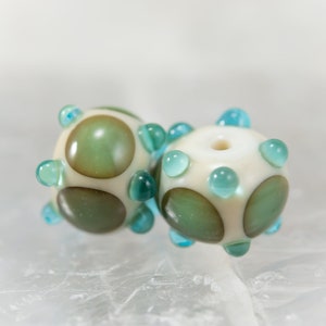 May include: Two handmade glass beads. Each bead is off-white with green and brown circular designs and teal-colored accents. The beads have a spherical shape with a hole through the center, and are suitable for jewelry making.