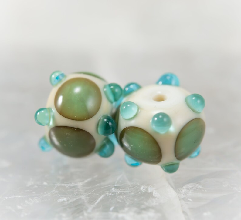 May include: Two handmade glass beads. Each bead is off-white with green and brown circular designs and teal-colored accents. The beads have a spherical shape with a hole through the center, and are suitable for jewelry making.