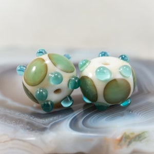 May include: Two handmade glass beads. Each bead is spherical, with a creamy white base color and large, irregular green and brown spots. Small, turquoise-colored glass accents are evenly spaced around the bead's surface. Each bead has a hole for stringing.